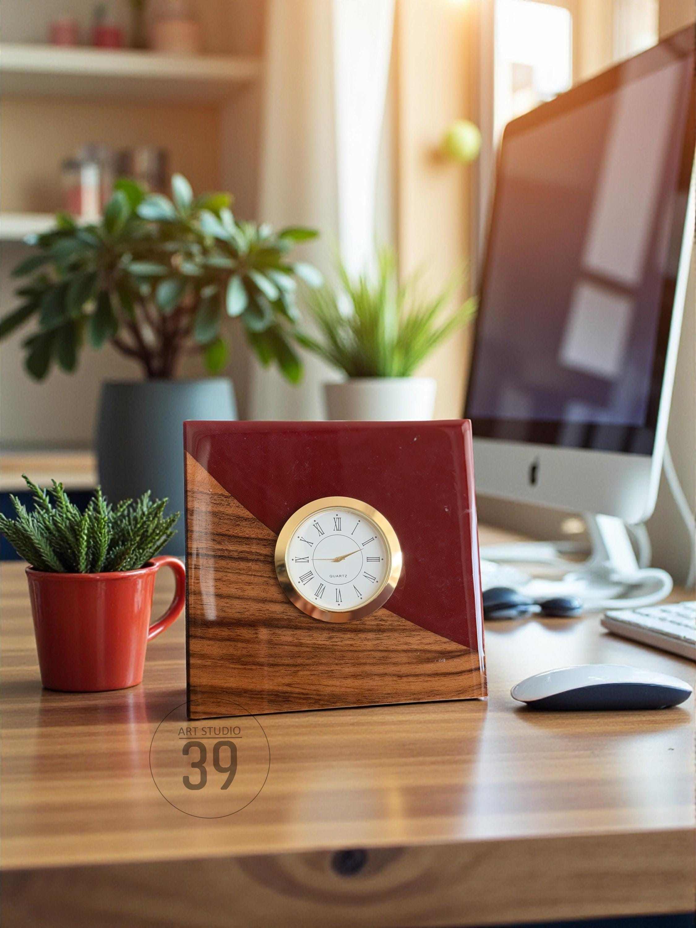 A wooden desk clock with a gold-colored bezel and white clock face. The clock has a red and wood grain design. A small red mug with a plant is to the left of the clock. The clock is on a wooden desk.