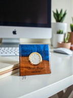A square desk clock with a gold-colored clock face and Roman numerals. The clock is set in a wooden base with a blue resin top. The text "Brooks Design" and "Liam Brooks" are engraved on the wood.