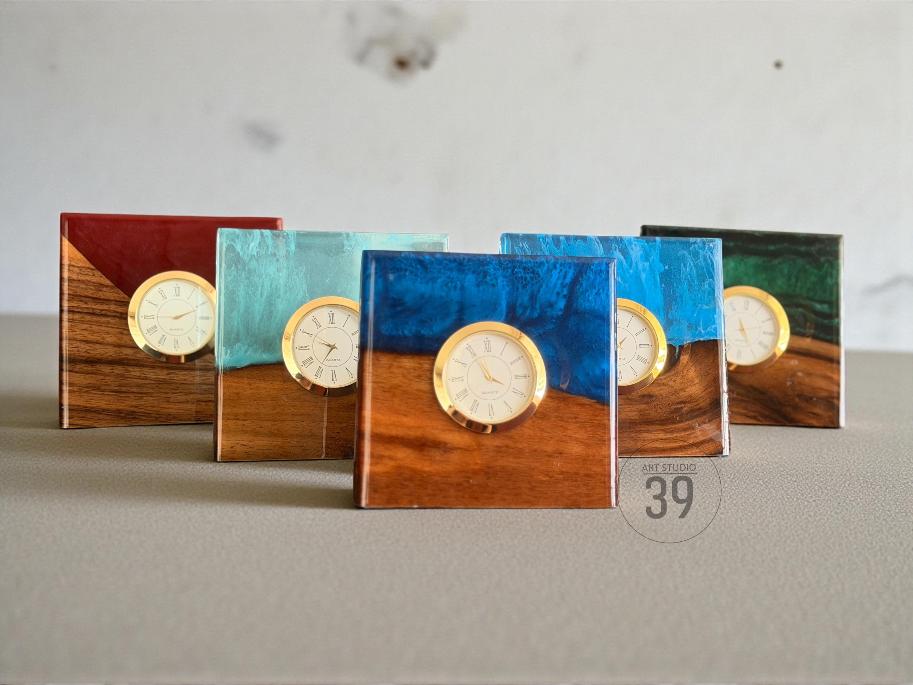A collection of square clocks with a wooden base and colorful resin tops. Each clock features a gold-colored frame and a white clock face. The resin colors include red, teal, blue, and green.