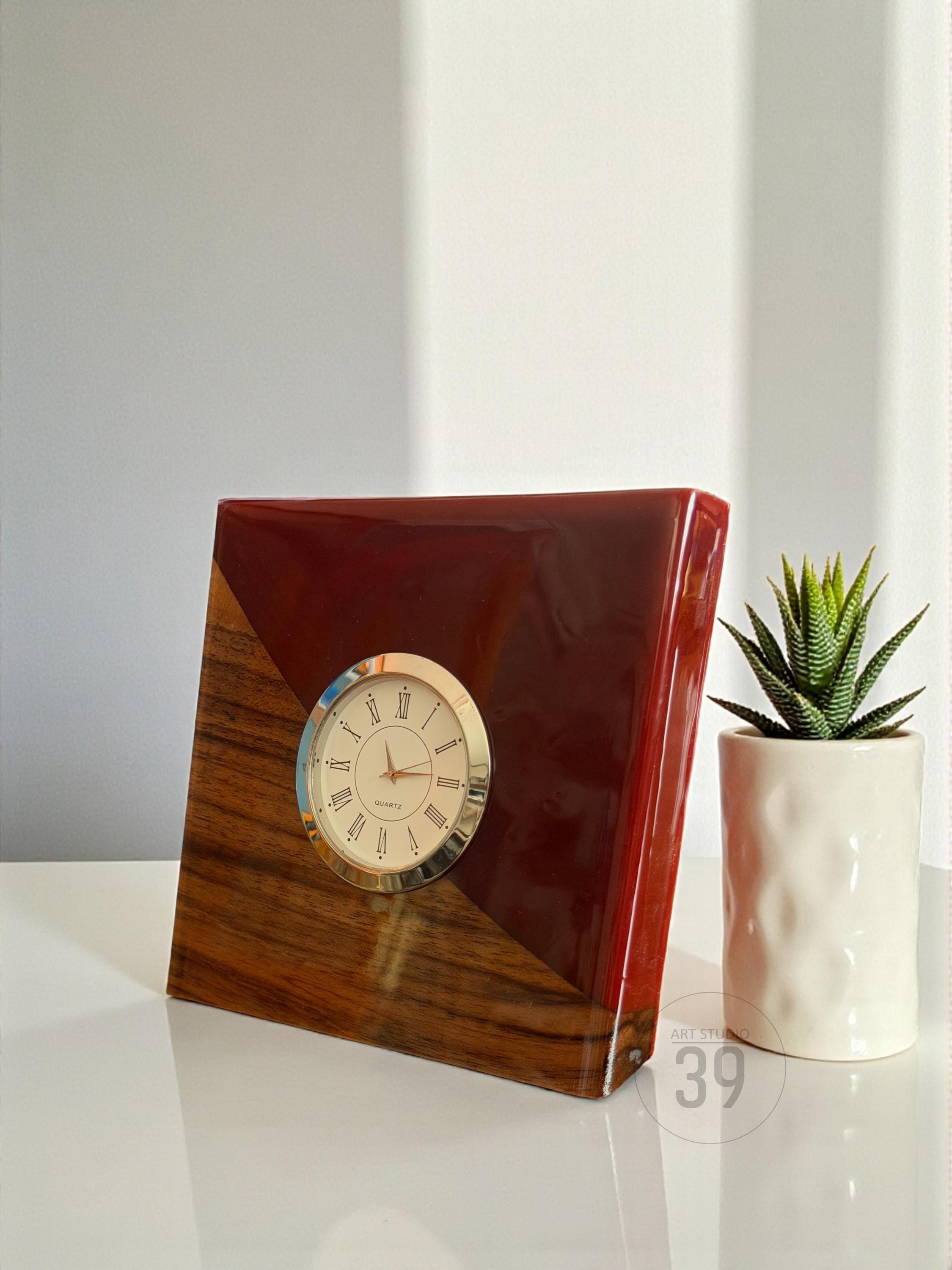 A square wooden clock with a dark red, glossy finish and a gold-colored clock face. The clock has a quartz movement and Roman numeral hour markers. The wooden base has a natural wood grain pattern. A small succulent plant in a white pot is next to the clock.