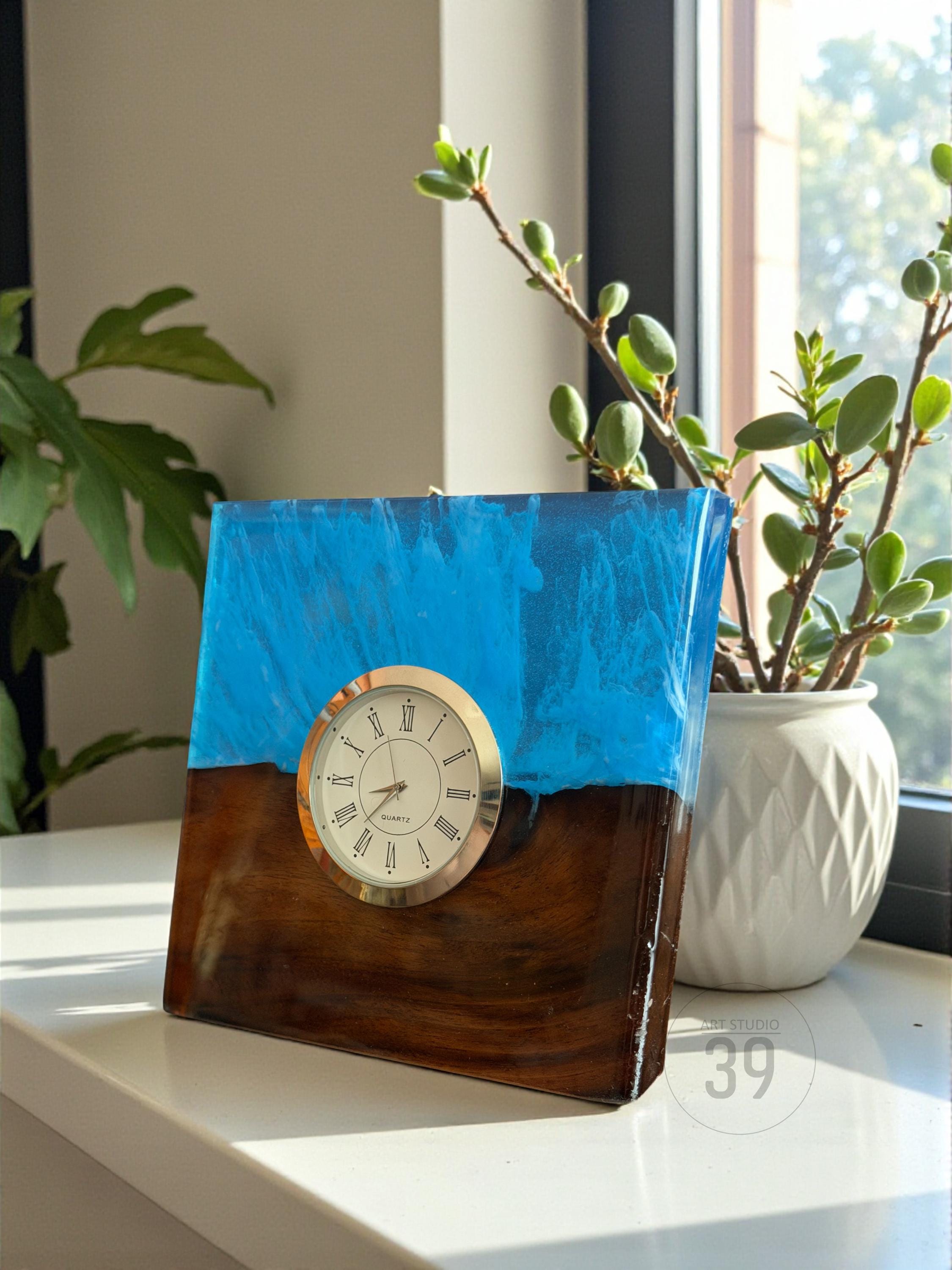 A square wooden clock with a blue resin top. The clock face is white with black Roman numerals and gold trim. The clock is sitting on a white surface near a window.