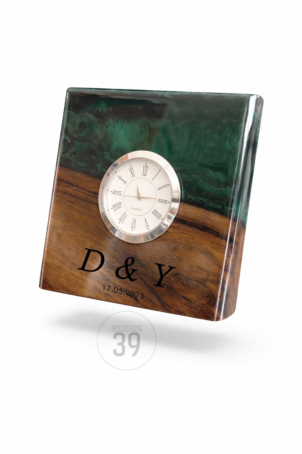 A square wooden clock with a silver-colored clock face. The top half is a dark green, swirled resin, and the bottom half is a brown wood grain. The clock is engraved with the letters "D &amp; Y" and the date "17.05.2023".