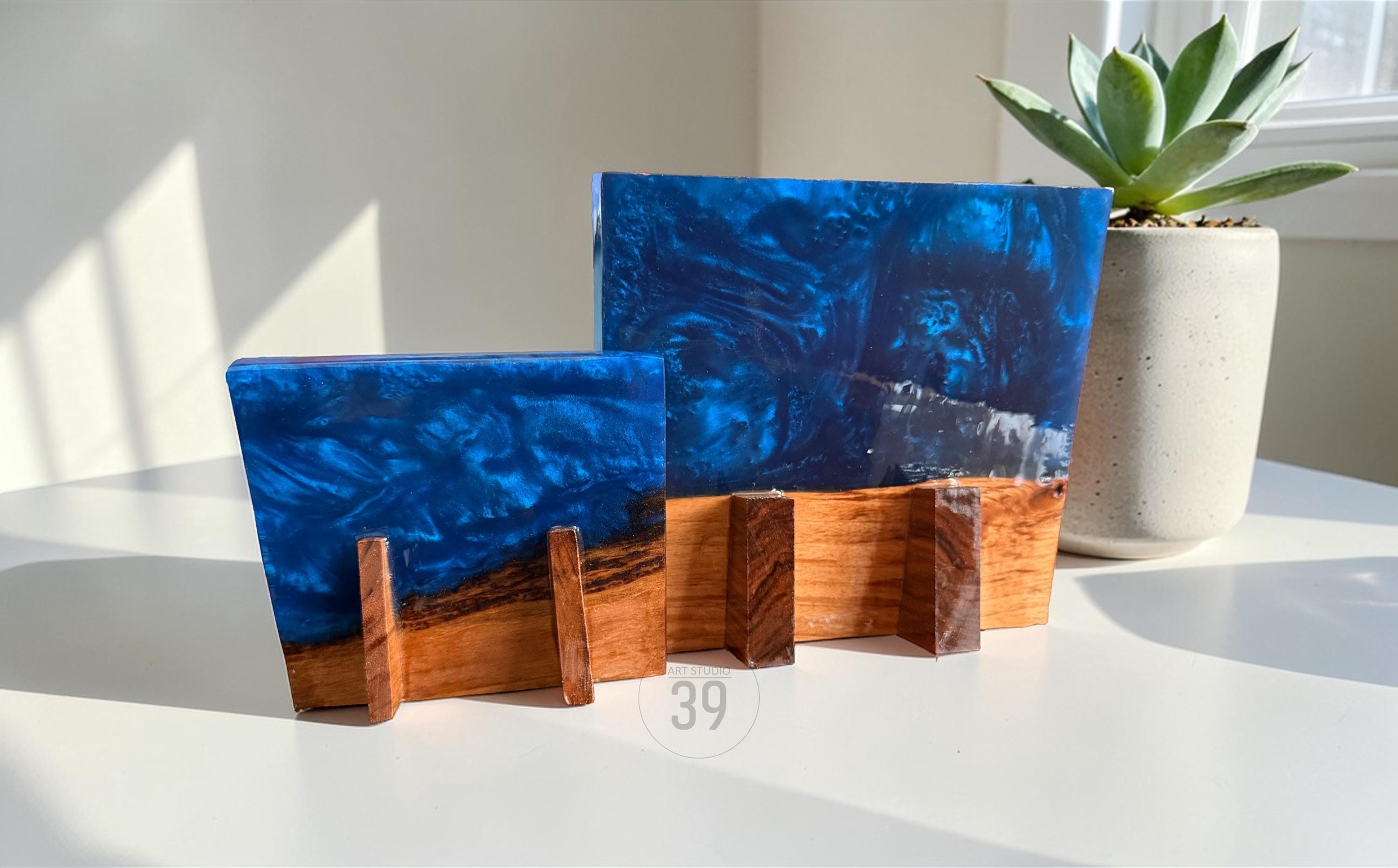 Two decorative resin art pieces with a blue ocean design, set on wooden stands. The resin has a swirling blue pattern with white accents, resembling waves. The wooden bases are a warm brown color. A succulent plant in a white pot is in the background.