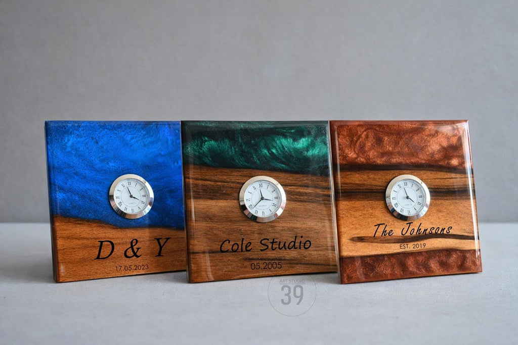 Three square wooden clocks with embedded resin and small clock faces. The clocks have different colors of resin: blue, green, and brown. Each clock has engraved text, including names and dates.