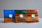Three square wooden clocks with embedded resin and small clock faces. The clocks have different colors of resin: blue, green, and brown. Each clock has engraved text, including names and dates.