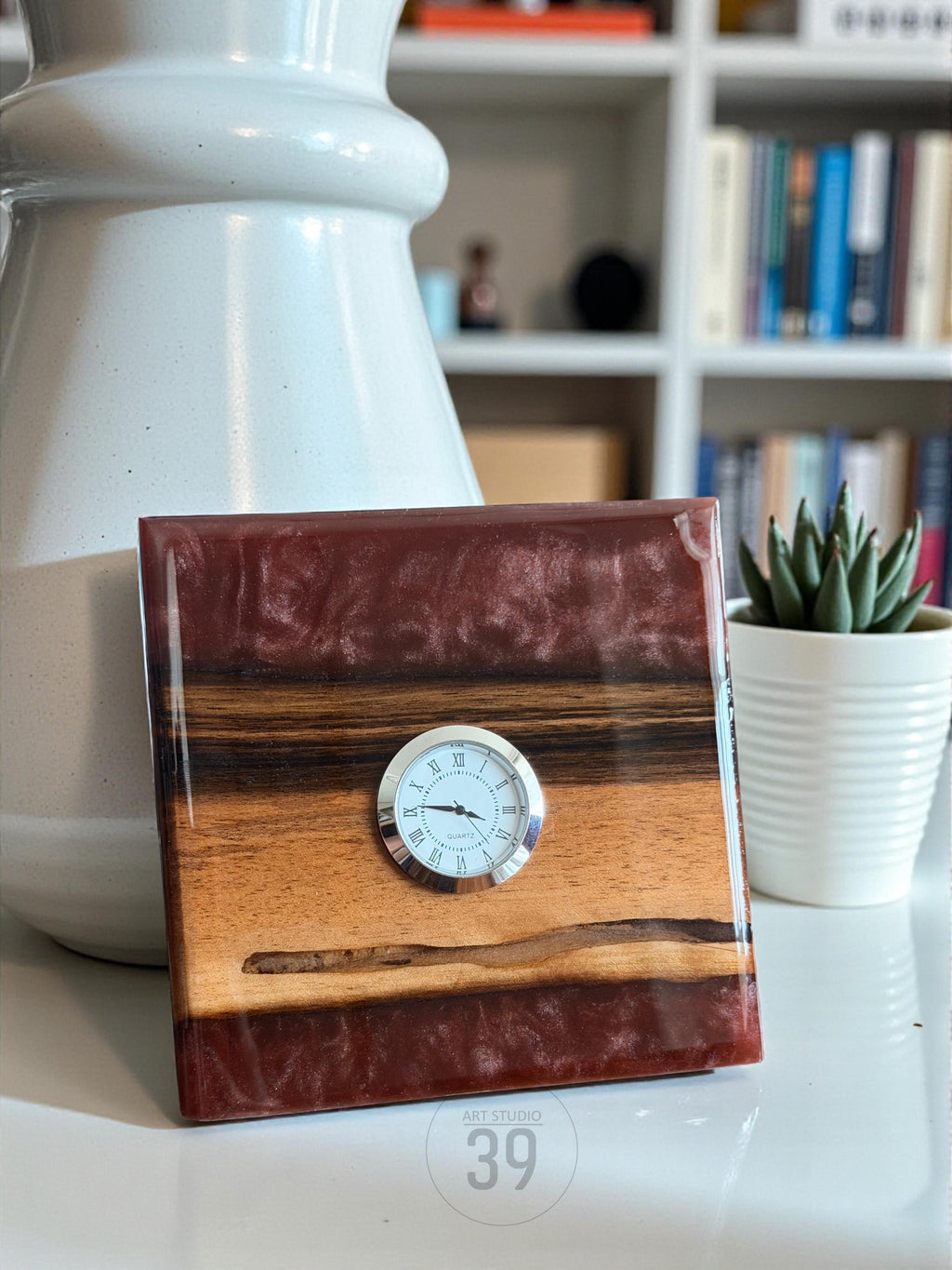 Square wooden clock with a silver-tone clock face and Roman numerals. The clock features a combination of wood and resin in shades of brown and burgundy. The text "ART STUDIO 39" is visible.