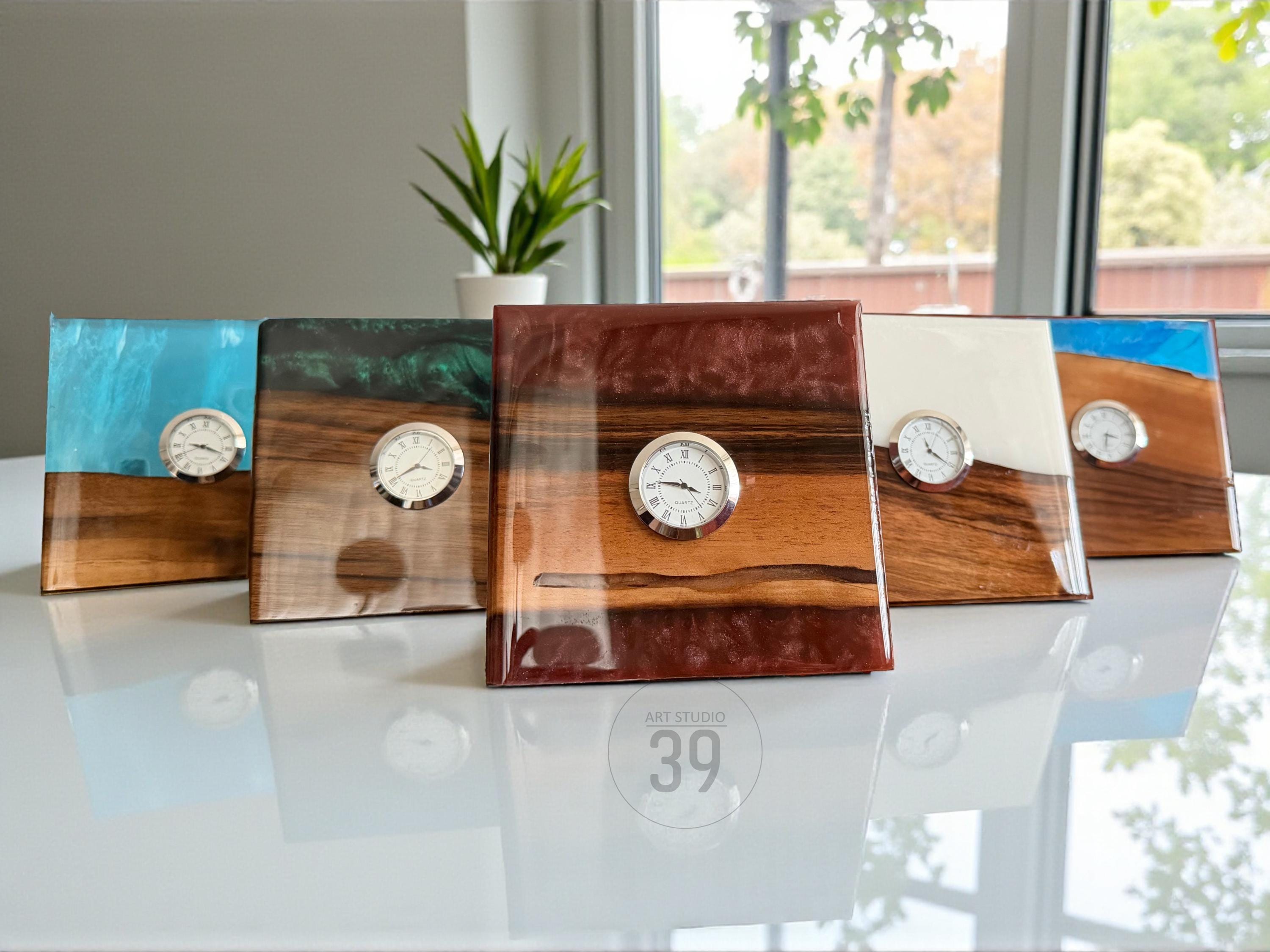 Five square wooden clocks with resin tops in various colors, including blue, green, white, and brown. Each clock features a silver-toned clock face. The clocks are displayed on a white surface.