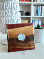 Square wooden clock with a silver clock face. The clock is made of wood and red resin. The clock face has black Roman numerals. The clock is on a white surface. The clock is a decorative item.
