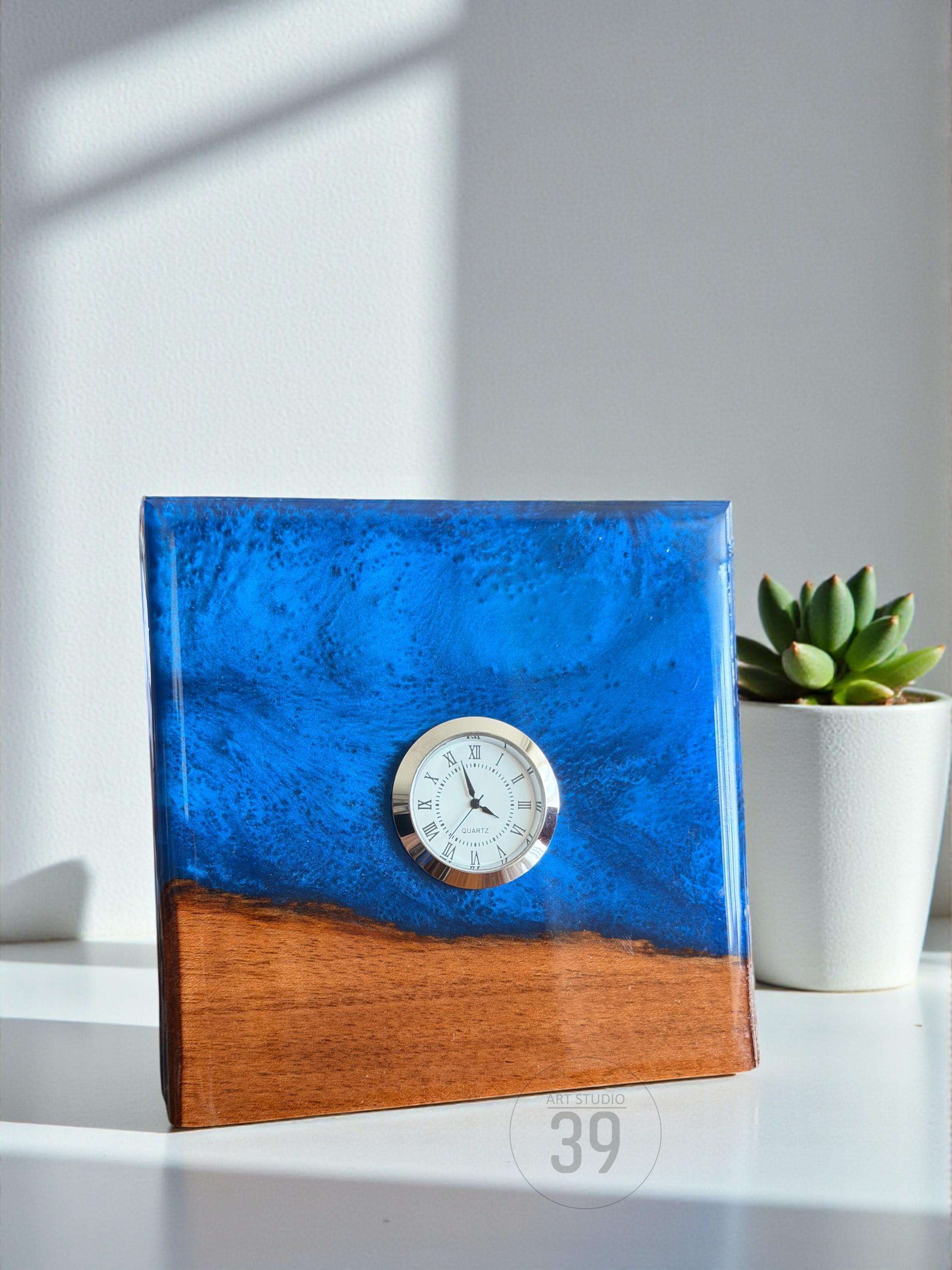 A square wooden clock with a blue resin top and a gold-rimmed clock face. The bottom half of the clock is a warm brown wood. A small succulent in a white pot sits to the right of the clock.