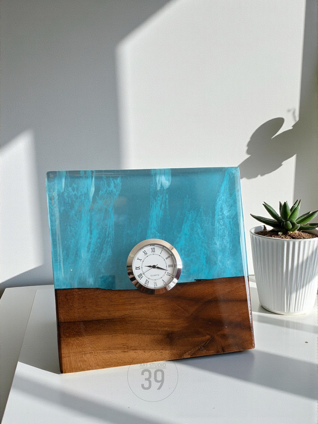 A square wooden clock with a turquoise resin top and a silver clock face. The bottom half is a rich brown wood. The clock has a classic Roman numeral clock face. A small succulent plant in a white pot is to the right.