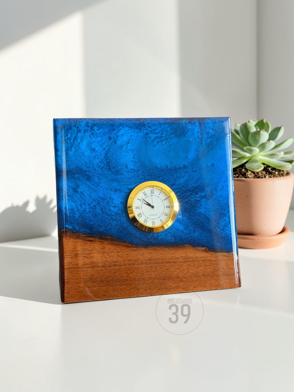 A square clock with a gold-colored clock face and Roman numerals. The top half is a vibrant blue resin, and the bottom is a warm brown wood. The clock is labeled "ART STUDIO 39". A succulent plant in a terracotta pot is in the background.