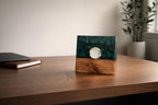 A square desk clock with a gold-tone bezel and white clock face. The top half is a dark green resin, and the bottom half is wood. The clock is on a wooden desk.
