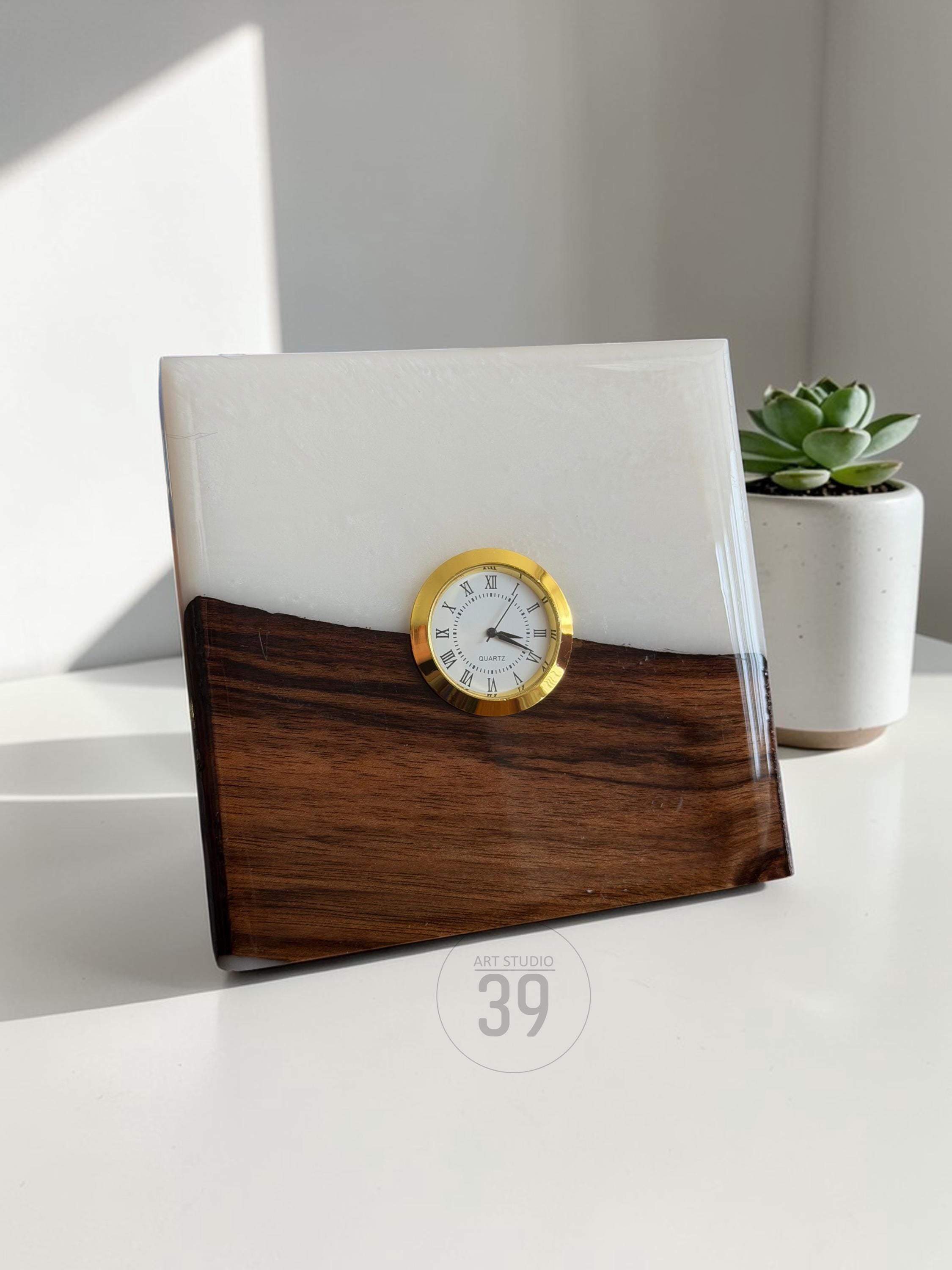 A square desk clock with a gold-tone clock face and Roman numerals. The clock is set in a block of wood and white resin. The bottom half is dark wood, and the top half is white. The clock has the text "ART STUDIO 39".