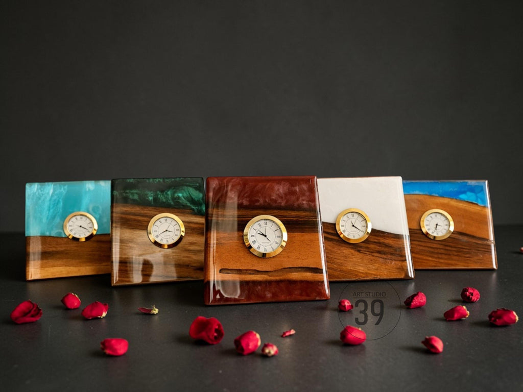 Five square wooden clocks with resin accents in blue, green, white, and brown. Each clock features a gold-colored frame and a white clock face. Red rose petals are scattered around the clocks.