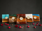 Five square wooden clocks with resin accents in blue, green, white, and brown. Each clock features a gold-colored frame and a white clock face. Red rose petals are scattered around the clocks.