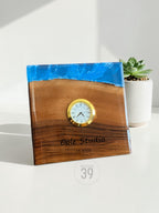 A square wooden clock with a gold-colored clock face. The top of the clock is a blue resin. The clock face has Roman numerals. The text "Cole Studio 05.2005" is engraved on the wood. The clock is labeled "ART STUDIO 39".