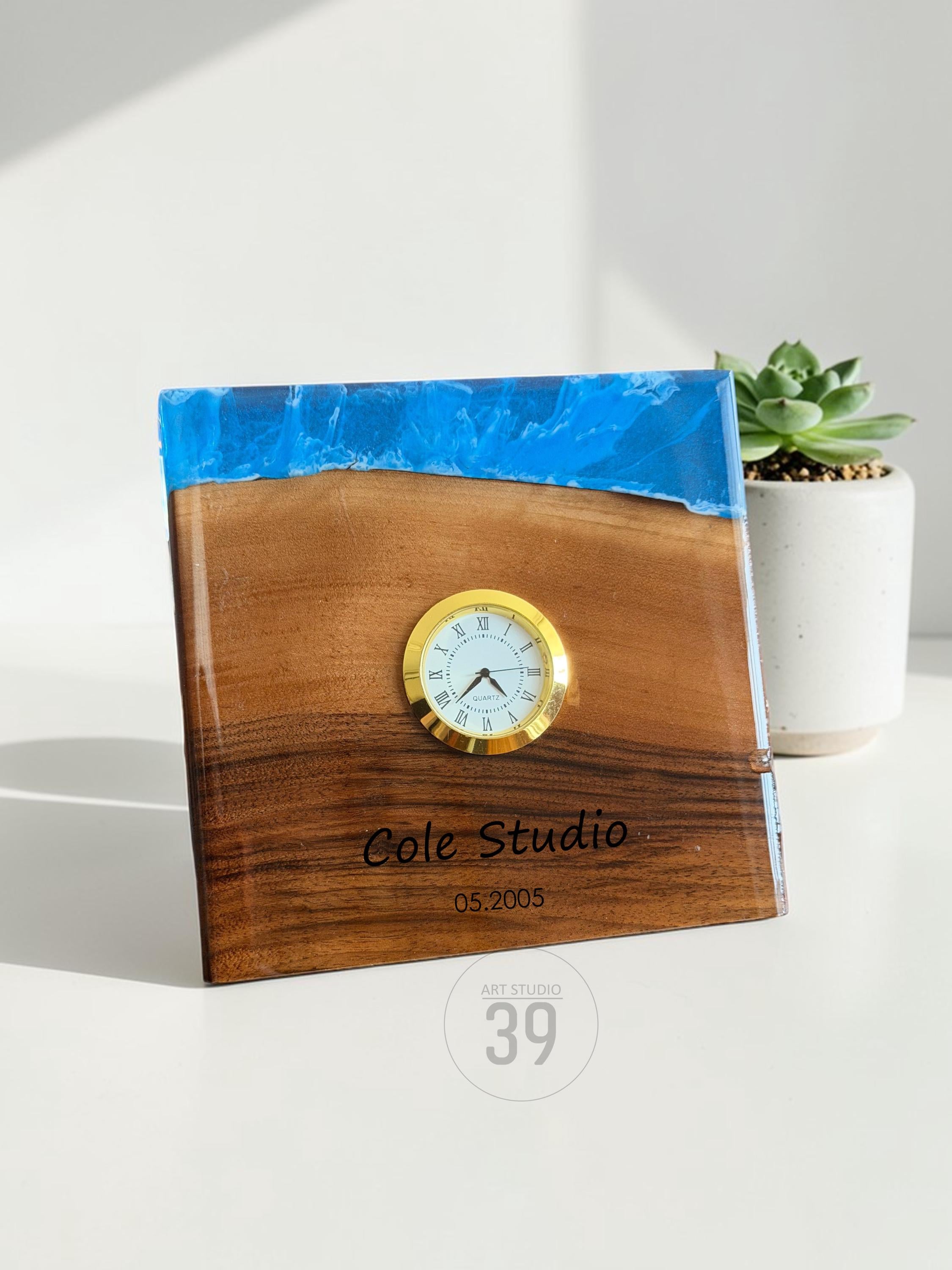 A square wooden clock with a gold-colored clock face. The top of the clock is a blue resin. The clock face has Roman numerals. The text "Cole Studio 05.2005" is engraved on the wood. The clock is labeled "ART STUDIO 39".