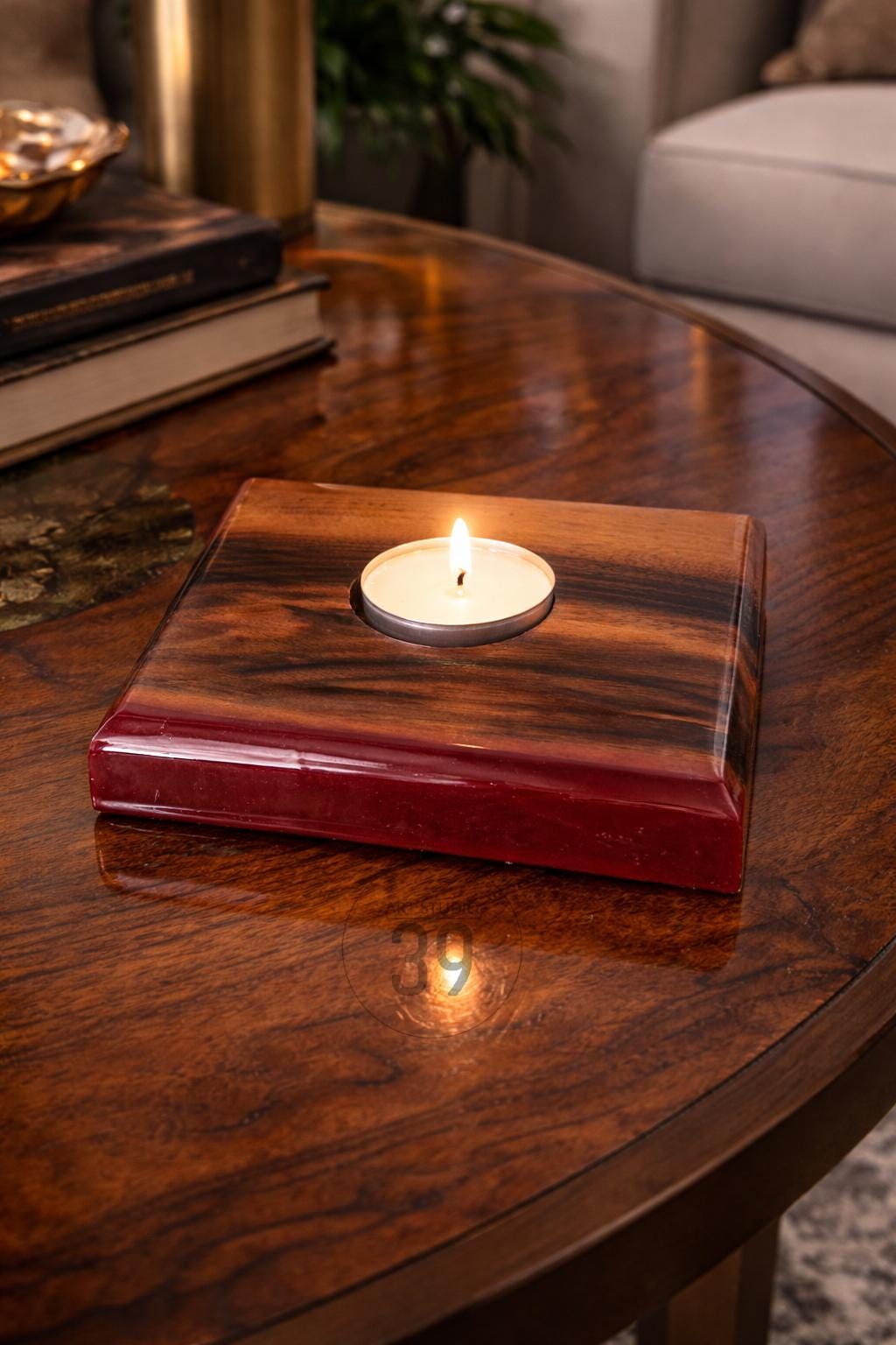 A square wooden candle holder with a lit tea light. The holder has a dark, patterned wood top and a red base. The candle flame is bright against the dark wood. The candle holder is on a wooden table.
