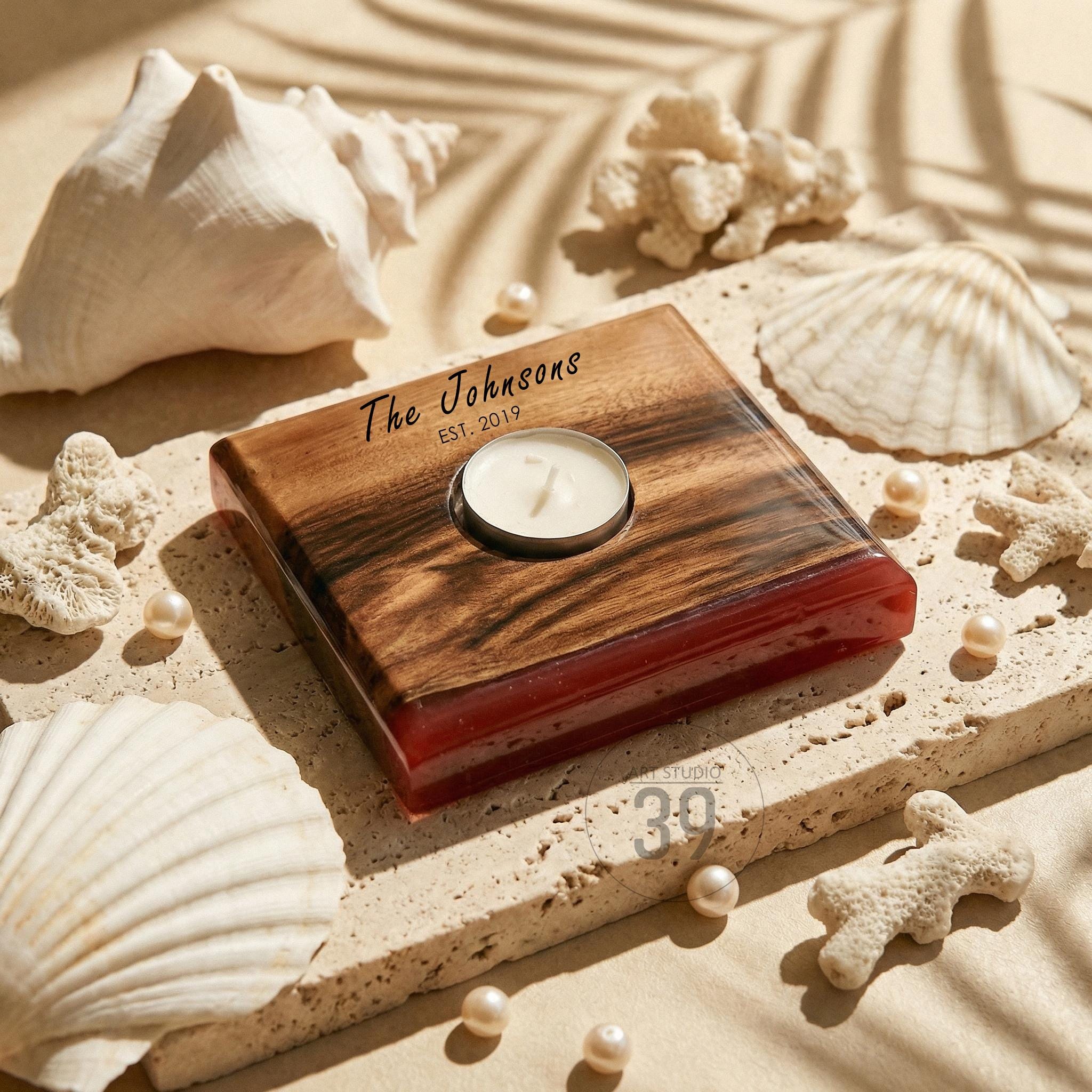 A wooden candle holder with a red base and a circular indentation for a tea light. The top is engraved with "The Johnsons EST. 2019". The holder is surrounded by seashells, pearls, and coral on a sandy surface.