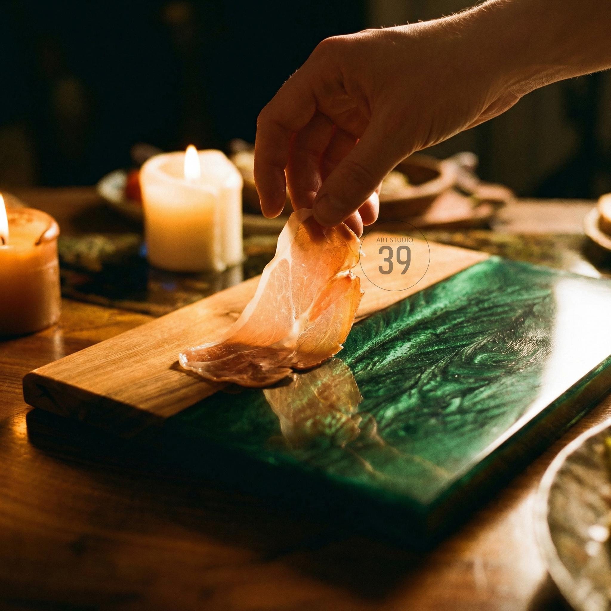 A wooden and green resin serving board with a slice of prosciutto. The board has a rectangular shape with a wooden section and a green, marbled resin section. The image includes lit candles and the text "ART STUDIO 39".