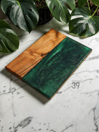 A rectangular wooden serving board with a section of deep green epoxy resin. The wood has a natural grain, and the resin has a swirling, glossy finish. The board is likely used for serving food or as a decorative item.