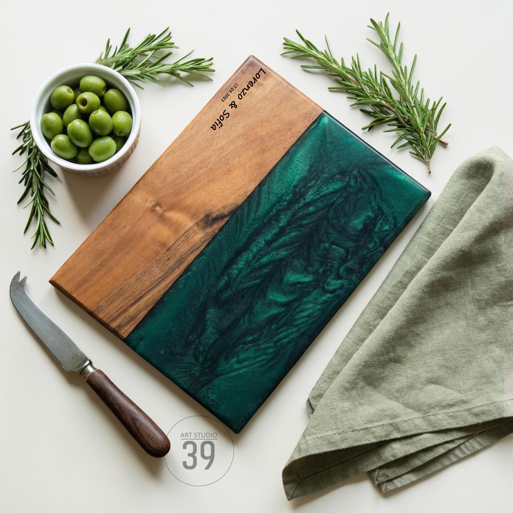 A rectangular wooden serving board with a dark green epoxy resin section. The board is accompanied by a cheese knife, a bowl of olives, rosemary sprigs, and a folded green linen napkin. The board is engraved with "Lorenzo &amp; Sofia" and a date.