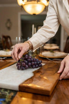 A rectangular serving board with a white and brown design. A bunch of dark purple grapes rests on the white section. The board is on a wooden table, and a person's hand is holding the grapes.
