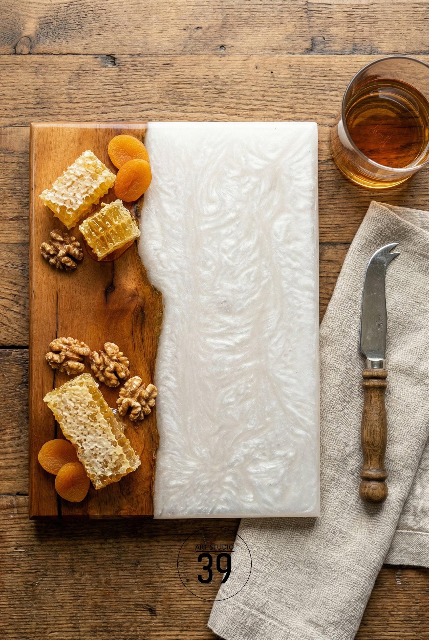A two-toned serving board with a wooden side and a white resin side. The wooden side holds honeycomb, walnuts, and dried apricots. A glass of amber liquid, a cheese knife, and a linen napkin are also present.