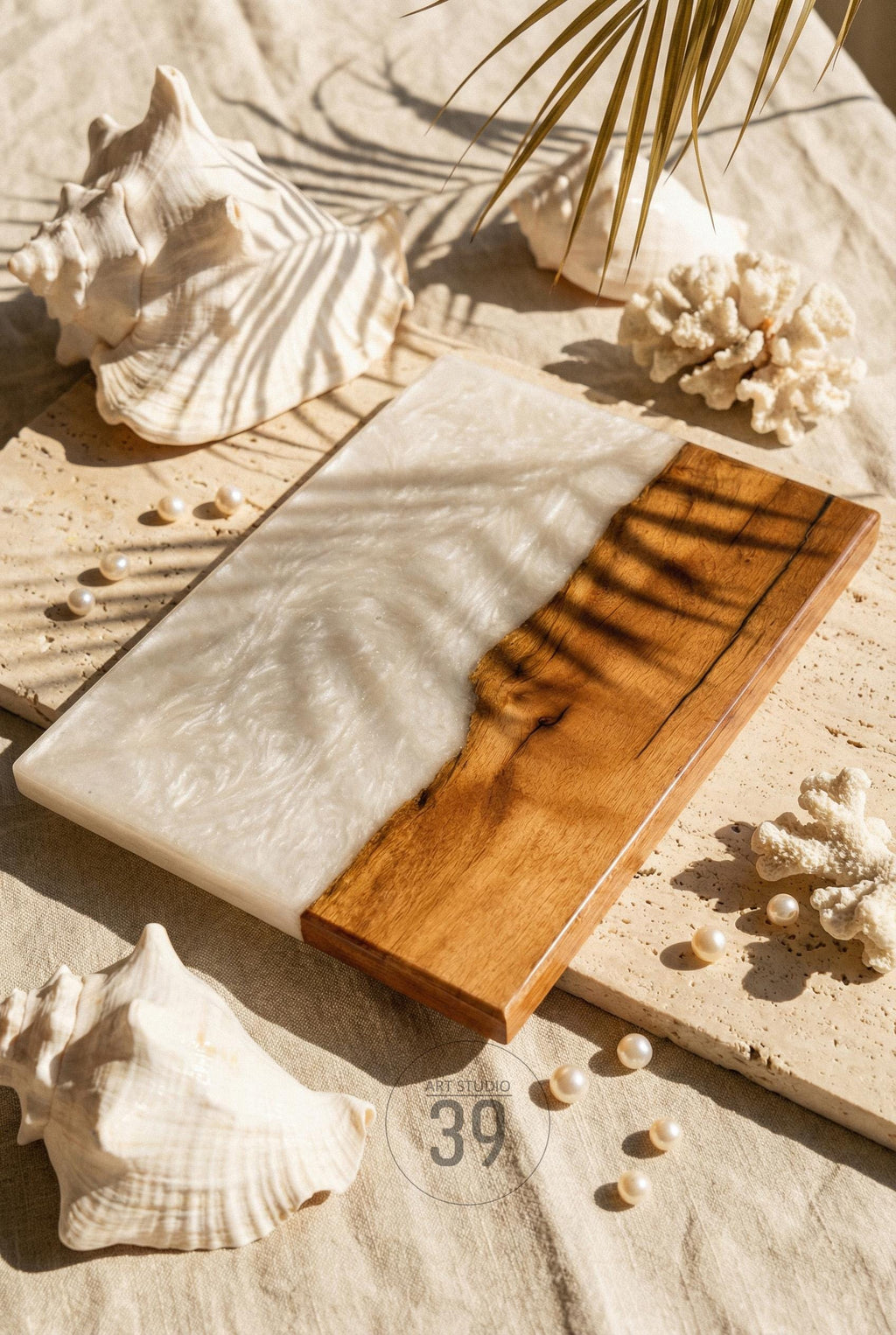 A rectangular serving board with a wood and white resin design. The wood portion is a warm brown, while the resin has a pearlescent white swirl. The board is surrounded by seashells, coral, and pearls.