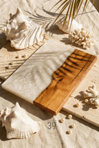 A rectangular serving board with a wood and white resin design. The wood portion is a warm brown, while the resin has a pearlescent white swirl. The board is surrounded by seashells, coral, and pearls.