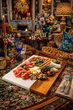 A charcuterie board with a white marble and wood design, laden with prosciutto, cheese, olives, nuts, and breadsticks. A blue glass and a lit candle are also on the table. The setting is a richly decorated room.