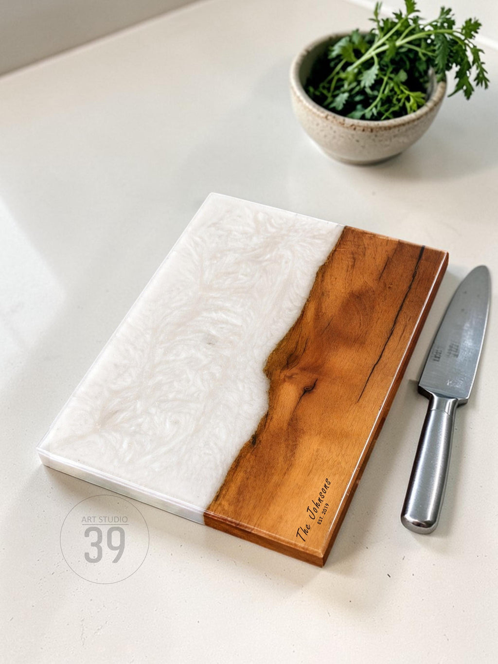 A rectangular cutting board with a white resin design on one side and natural wood on the other. The wood side has engraved text. A small knife rests beside the board. A small bowl of green herbs is in the background.