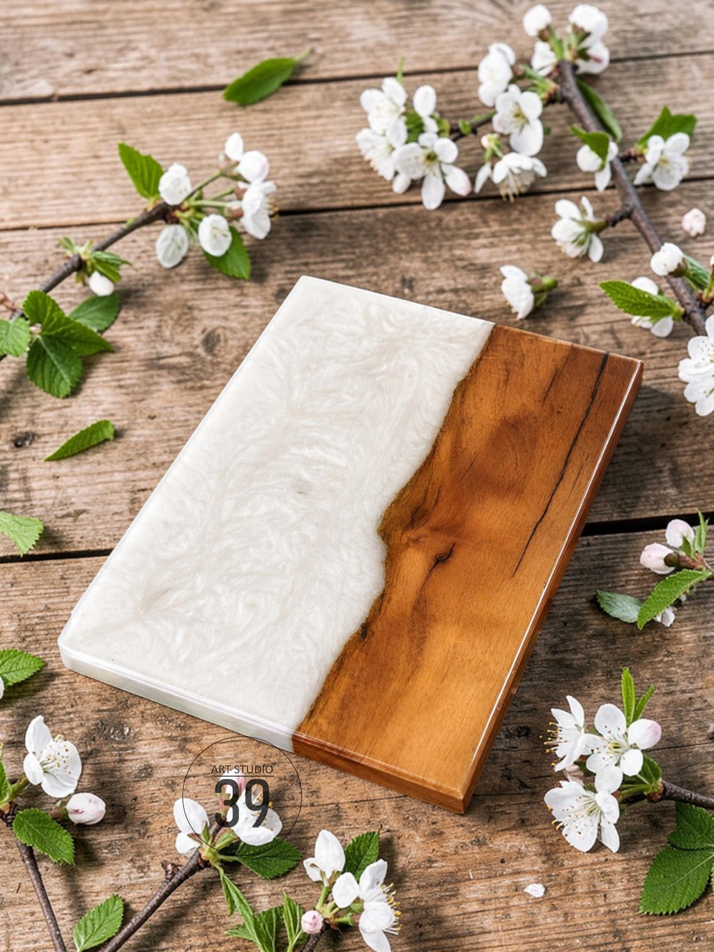 A rectangular serving board with a two-tone design. One half is a glossy, swirling white resin, and the other is a polished, warm-toned wood. The board is surrounded by white blossoms and green leaves on a wooden surface.