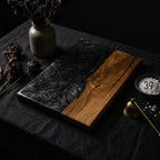 A rectangular wooden serving board with a black resin design on one side and natural wood grain on the other. The board is on a dark surface, with a small bell and a dish with the number 39 visible.