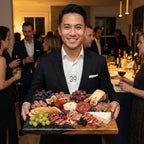 A man in a black suit holds a wooden charcuterie board with an assortment of cheeses, meats, crackers, grapes, figs, and berries. The board has a dark marble-like edge. The man is smiling at the camera. The text "ART STUDIO 39" is on his shirt.