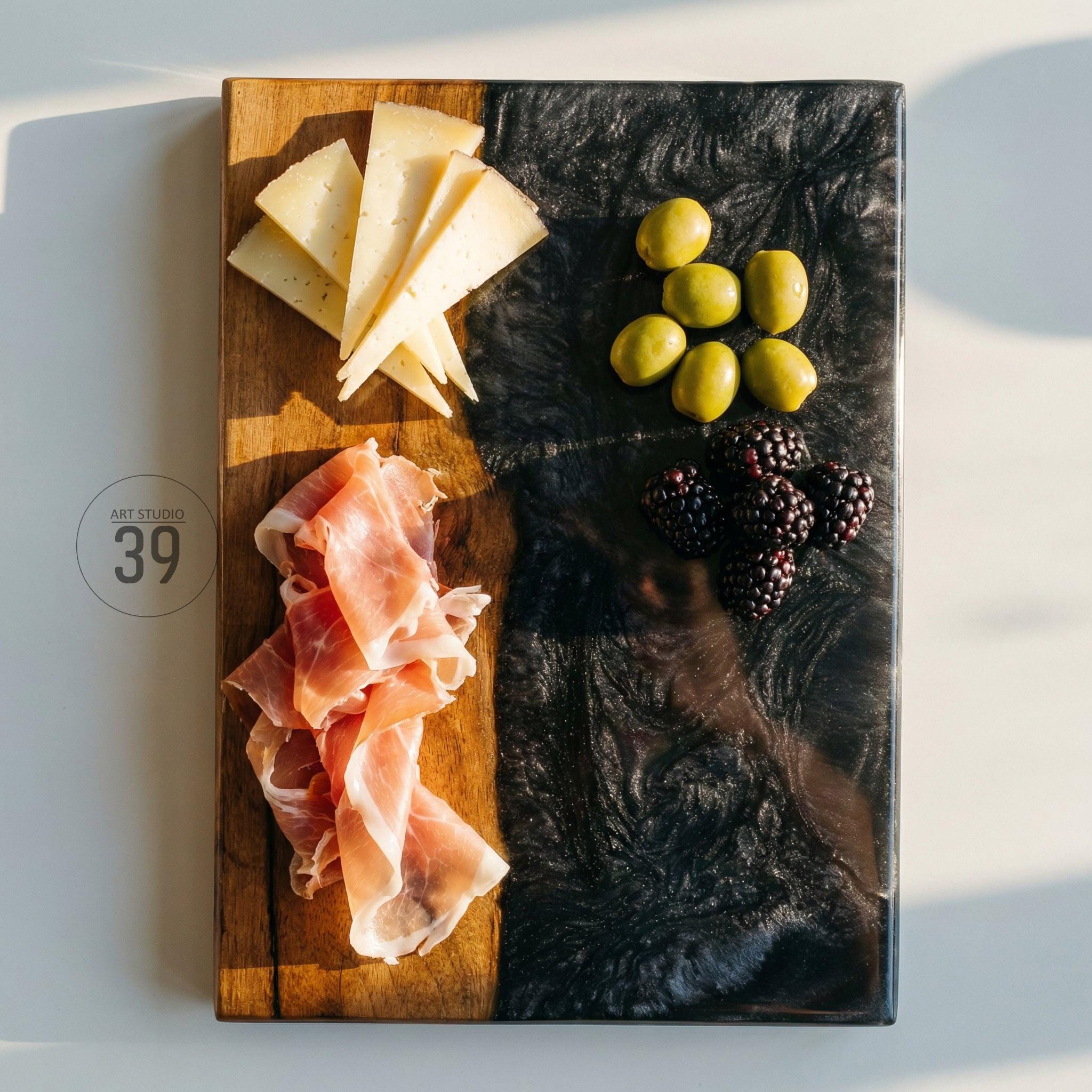A rectangular wooden serving board with a black resin section. The board is laden with sliced cheese, prosciutto, green olives, and blackberries. The text "ART STUDIO 39" is printed on the board.