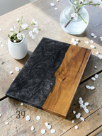 A rectangular serving board with a striking design. One half features a swirling black epoxy resin, while the other showcases natural wood grain. The board is branded with "ART STUDIO 39". Decorative white flowers and vases are in the background.