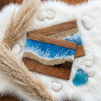 Two wooden serving boards with a resin ocean design. The boards feature a dark wood frame, a sandy beach, and a vibrant blue ocean with white waves. The boards are surrounded by white stones and fluffy beige pampas grass.