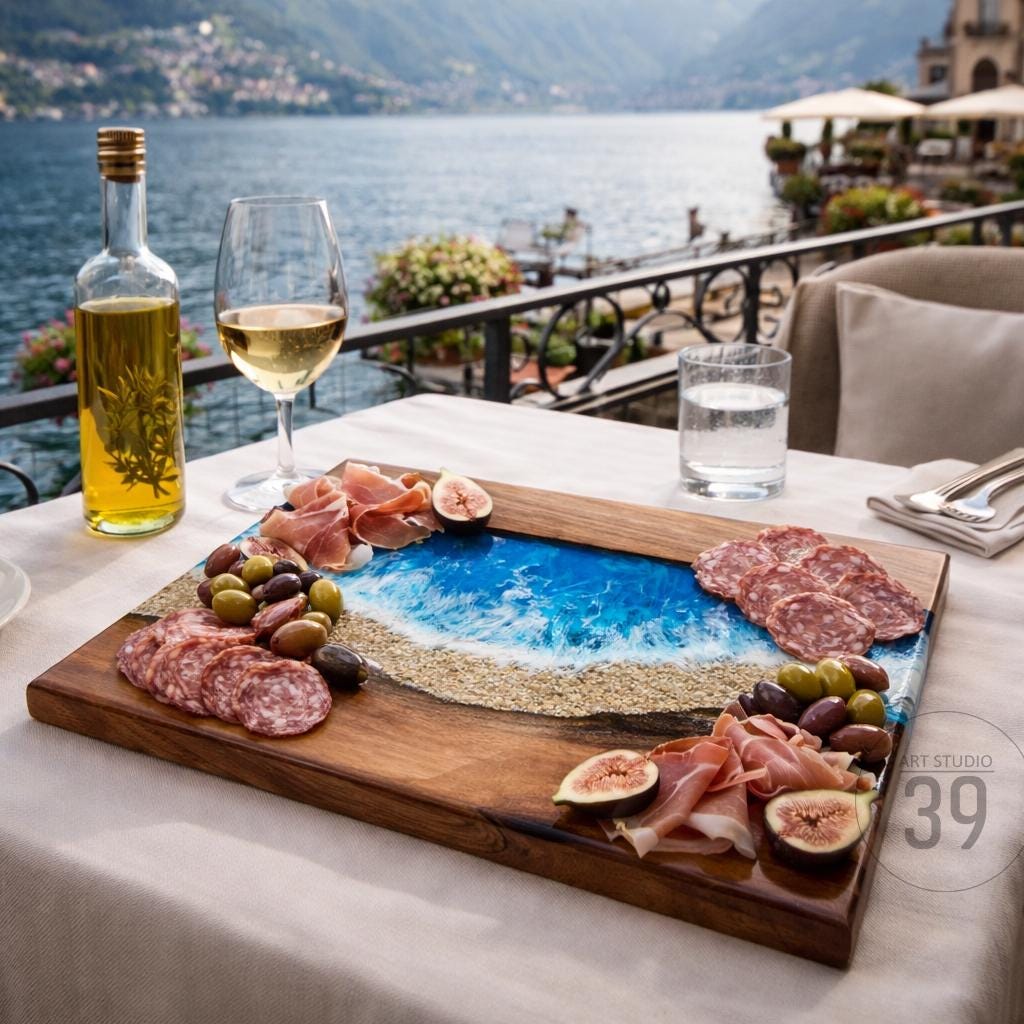 A wooden serving board with a blue and sand-colored resin design resembling a beach scene. The board is laden with olives, salami, prosciutto, and figs, alongside a glass of wine and a bottle of oil.