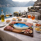 A wooden charcuterie board with a resin ocean design, filled with olives, meats, and figs. The board is on a white tablecloth, surrounded by wine glasses and small bowls of snacks. The background features a lake and mountains.