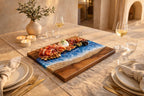 A wooden serving board with a resin design resembling a beach scene, featuring blue waves and a sandy shore. The board is laden with food items like prosciutto, figs, and olives. Wine glasses and candles are also present.