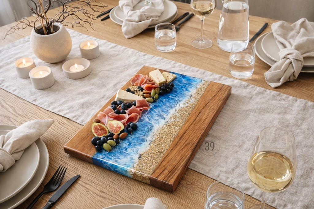 A wooden serving board with a blue resin wave design and a sandy beach effect. The board is laden with food items like cheese, prosciutto, figs, and grapes. The scene is set on a wooden table with candles and glassware.