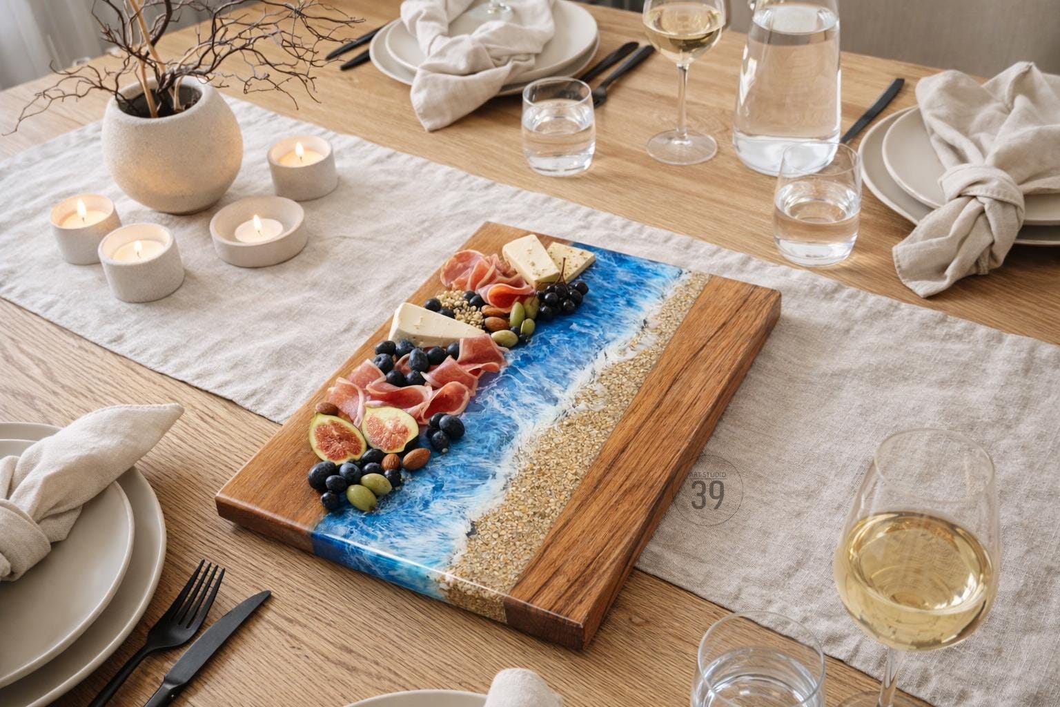 A wooden serving board with a blue resin wave design and a sandy beach effect. The board is laden with food items like cheese, prosciutto, figs, and grapes. The scene is set on a wooden table with candles and glassware.