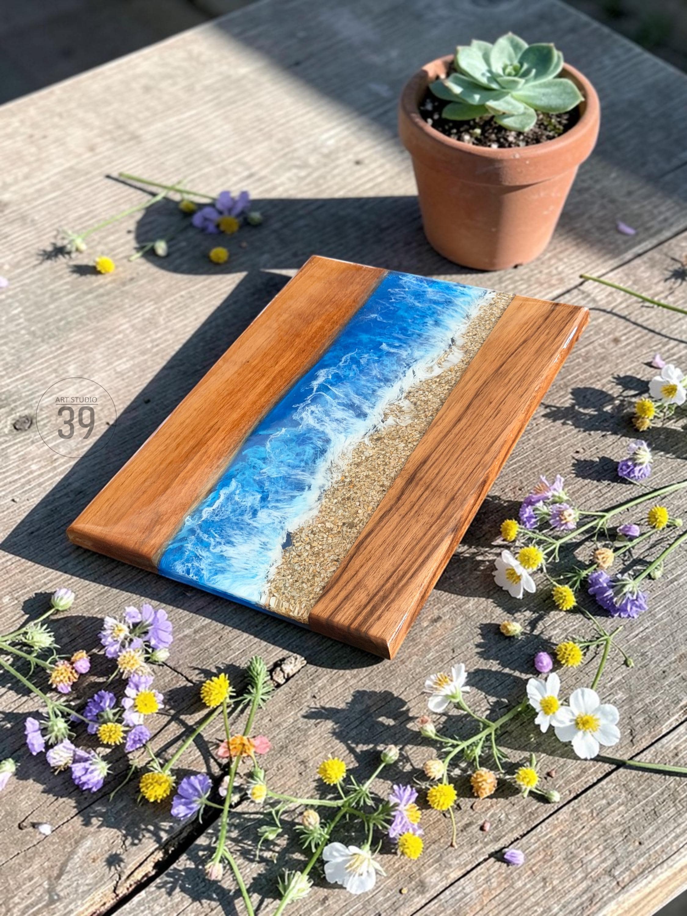 Wooden serving board with a beach scene design. The board features a central section of sand and a blue resin ocean with white foamy waves. The board is surrounded by natural wood and decorated with flowers.