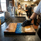 A wooden cutting board with a blue resin river design. The board has a natural wood grain on either side of the blue resin, with a sandy beach-like strip. The board is being cleaned by a person wearing black gloves.