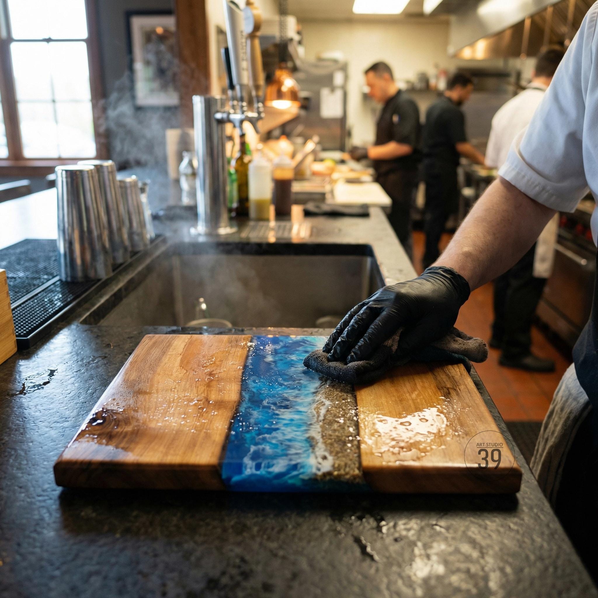 A wooden cutting board with a blue resin river design. The board has a natural wood grain on either side of the blue resin, with a sandy beach-like strip. The board is being cleaned by a person wearing black gloves.