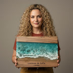 A wooden framed resin art piece depicting an ocean scene with turquoise waves and sandy shore. The artwork includes the names "Lorenzo &amp; Sofia" and the date "17.05.2023". The frame is made of wood.
