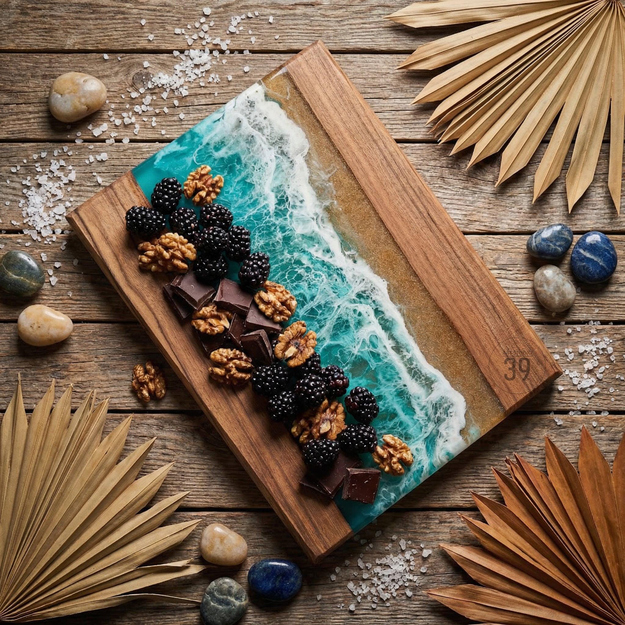 A wooden serving board with a resin ocean design, adorned with blackberries, walnuts, and chocolate. The board has a natural wood grain and is decorated with small stones and dried palm leaves.