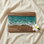 Wooden framed artwork featuring a resin ocean scene with turquoise waves and sandy shore. The text "The Johnsons EST. 2019" is displayed on the wooden frame. A white flower rests on the right side of the artwork.
