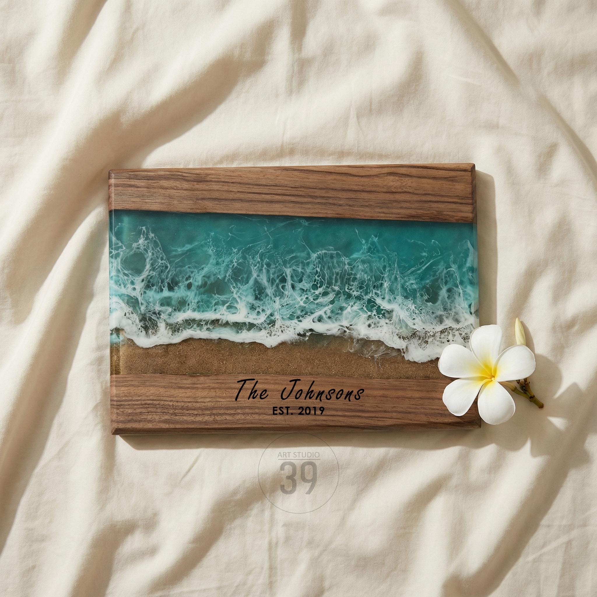 Wooden framed artwork featuring a resin ocean scene with turquoise waves and sandy shore. The text "The Johnsons EST. 2019" is displayed on the wooden frame. A white flower rests on the right side of the artwork.